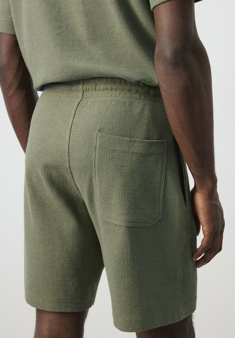 Homme portant un short en maille vert olive avec une poche patch à l'arrière et un haut assorti, vu de dos.