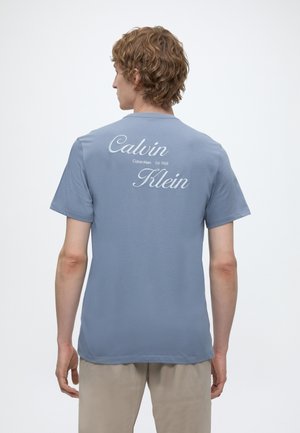 Göndör hajú férfi, kék Calvin Klein pólót visel logóval és az „Est. 1968” felirattal a hátán, bézs nadrággal párosítva.