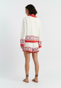 Witte gebreide twee-delige outfit met rode en blauwe bloemenpatronen, bestaande uit een boxy cardigan en shorts. Korte mouwen met ribbels.