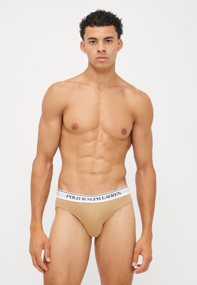 Beige férfi alsónadrág fehér derékszíjjal, amelyen fekete "POLO RALPH LAUREN" felirat található; sima textúrájú és testhez simuló kialakítású.