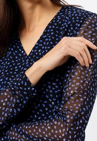 Top trasparente blu navy con scollatura a V, maniche lunghe e motivo a pois blu. I polsini hanno dettagli con bottoni; il tessuto appare leggero e strutturato.