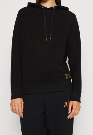 Osoba ubrana w czarną bluzę z kapturem na długim rękawie z troczkami oraz czarne spodnie z haftowanymi na złoto inicjałami "A|".