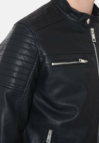Giacca da motociclista nera in pelle con dettagli a spina sulle spalle, tasche con zip argentate e colletto a scatto. Texture liscia e design aderente.