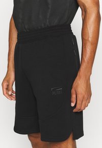 Černé sportovní kraťasy z měkké tkaniny, se žebrovaným pasem, bočními kapsami a vyraženým logem Puma na spodní části přední strany.