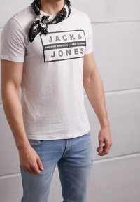 White short-sleeve t-shirt featuring black text "JACK&JONES." Worn with a black and white patterned bandana around the neck. Light denim jeans.