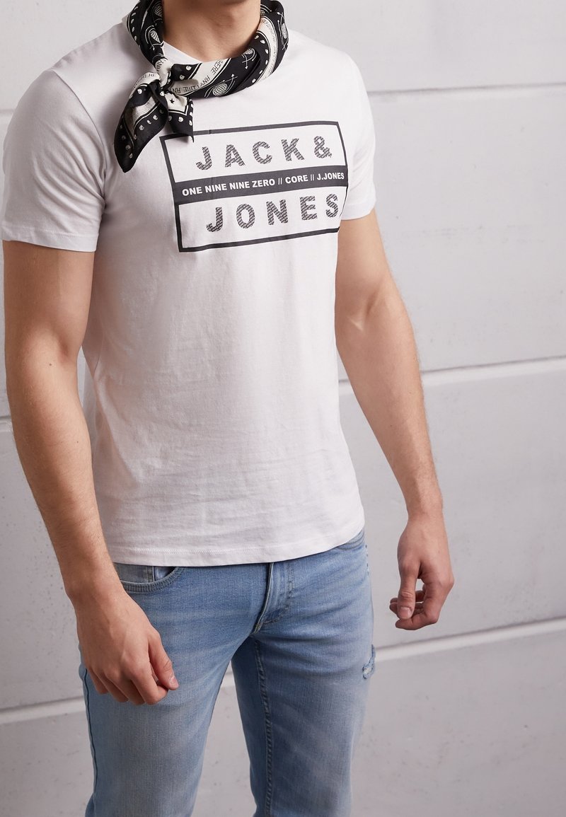 White short-sleeve t-shirt featuring black text "JACK&JONES." Worn with a black and white patterned bandana around the neck. Light denim jeans.