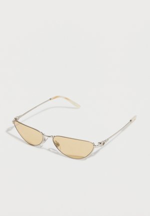 Lunettes de soleil à monture argentée fine, avec des verres étroits teintés jaune et des branches beige incurvées, posées à plat sur une surface blanche.