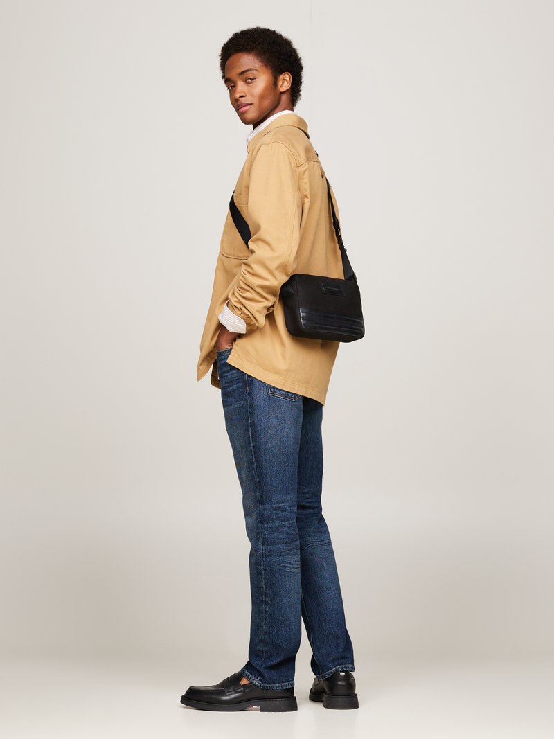 Borsa a tracolla nera con superficie texture, indossata sopra una camicia button-up beige, abbinata a jeans blu e mocassini neri.