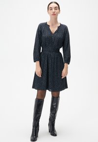 Dunkelblaues Blumenkleid mit Dreiviertelärmeln, gerafftem Taillenschnitt und ausgestelltem Rock, kombiniert mit hohen schwarzen Lederstiefeln, vor einem schlichten Hintergrund.