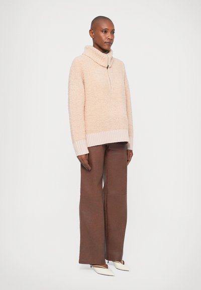 Maglione rosa chiaro, morbido, con colletto alto e mezza zip. Abbinato a pantaloni larghi marroni e scarpe bianche a punta. Design semplice.