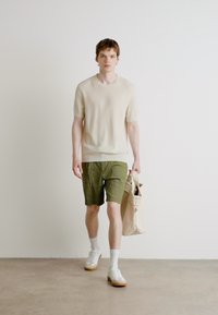 Maglione corto a maniche corte a costine crema, pantaloni corti verde oliva, sneakers bianche e borsa a tracolla beige chiara. Abbigliamento semplice e colori neutri.