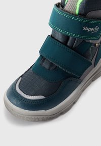 Chaussure montante teal et grise avec des bandes auto-agrippantes, tissu texturé et semelle en caoutchouc. Dispose d'un capuchon de pied lisse et de coutures contrastantes.