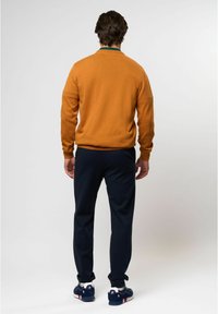 Homme vu de dos portant un pull moutarde, un pantalon bleu marine et des baskets bleu marine avec des semelles blanches sur un fond uni.