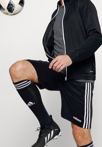 Schwarze Trainingsjacke mit weißem Reißverschluss, graues T-Shirt, schwarze Shorts mit weißen Streifen, schwarze Socken und schwarze Fußballschuhe. Ein Fußball ist in Bewegung.