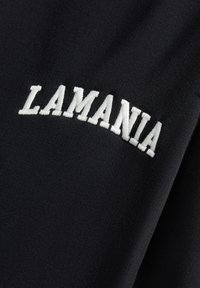 Juodas audinys su balta siuvinėta užrašu "LAMANIA" drąsiais, šiek tiek išlenktais didžiosiomis raidėmis.