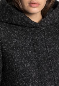 Cappotto di lana grigio scuro con una superficie texture, dotato di un ampio cappuccio, cuciture visibili e una vestibilità leggermente oversize.