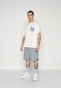 Bijela pamučna majica s ljubičastim grafičkim džepom, uparena sa svijetlosivim šortsevima. Uz to bijele tenisice i sive čarape do sredine lista.