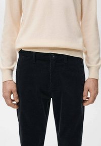 Hellbeiger Pullover über dunkelbraunen Cordhosen. Die Hose hat eine vertikal gerippte Textur und einen Knopfverschluss an der Taille.