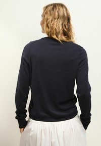 Marineblauwe gebreide trui met lange mouwen, ronde halslijn en geribbelde manchetten. Glad textuur, aansluitend op de taille, gedragen met een witte rok.