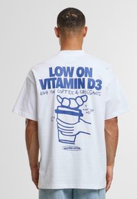 Valkoinen puuvillapaita, jossa on sininen graafinen teksti ja kuvia kädestä, joka pitää kahvikuppia, jonka päällä on croissantteja. Rentouduva istuvuus.