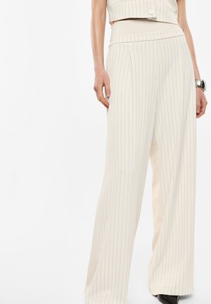 Donna che indossa pantaloni crema a vita alta e a gamba larga con sottili righe verticali e un top corto abbinato, accessoriata con un braccialetto d'argento.