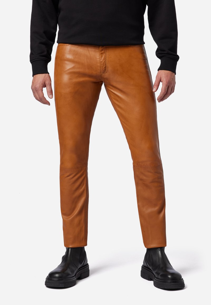 Bruine leren broek met een nauwsluitend ontwerp, voorzien van een gladde textuur en een middelhoge taille. Het model draagt ze in combinatie met zwarte enkellaarzen.