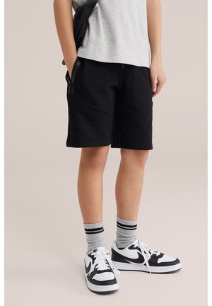 Persona vistiendo pantalones cortos negros, camiseta gris, calcetines grises con rayas negras y zapatillas Nike en blanco y negro, de pie con las manos en los bolsillos.