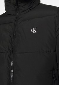Svart puffjacka med hög krage och hel dragkedja. Framträdande vit "cK"-logga på övre vänstra sidan. Slät, quiltad textur.