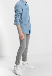 Camisa de botones azul claro en una tela suave, combinada con pantalones grises ajustados. Zapatillas blancas con acentos azules y puños enrollados completan el look.