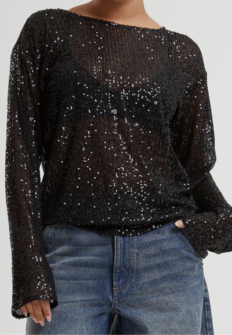 Haut noir transparent à manches longues, orné de petits sequins noirs, porté avec un jean en denim bleu, main dans la poche.