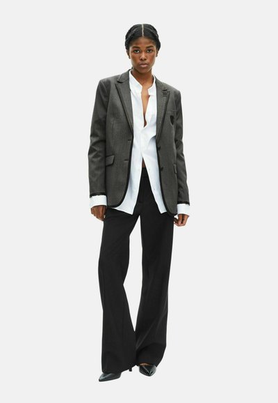Grijze stropdas-blazer, witte blouse en zwarte wijde broek. Soepele texturen, scherpe kraag en op maat gemaakte pasvorm. Zwarte puntige schoenen.