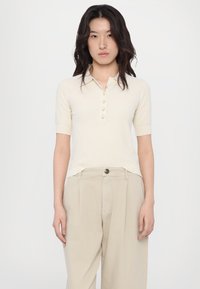 Short-sleeve beige polo shirt with a textured fabric, five button placket, and a collared neckline, paired with light beige trousers.