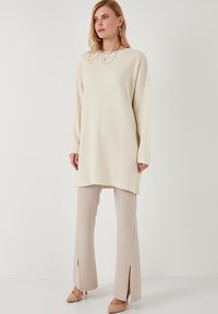 Tunique en tricot côtelé crème avec une coupe surdimensionnée, associée à un pantalon beige à fente haute et des talons nus. Accents de bijoux simples.