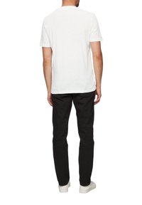 Witte katoenen T-shirt met korte mouwen, een ronde halslijn en een iets langere achterkant, gecombineerd met zwarte broeken en witte sneakers.
