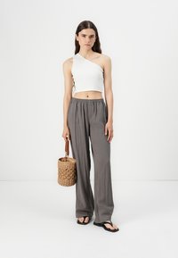 Haut court blanc à épaule dénudée, pantalon gris à jambes larges et tongs noires. Tient un sac en paille tressée avec une base ronde et une bandoulière.