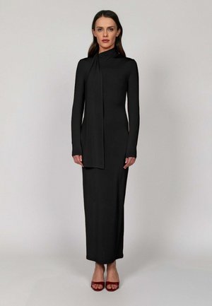 Kobieta w długiej czarnej dopasowanej sukience z wysokim kołnierzem i drapowanym detalem, w czerwonych butach na obcasie z odkrytymi palcami, stojąca na jednolitym tle.