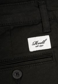 Material de mezclilla negra con un bolsillo cosido, que presenta una etiqueta blanca con la inscripción "Reell est. 1997" y un detalle de botón negro.