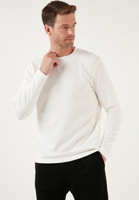 Weißes Langarmhemd mit strukturiertem, geripptem Design. Rundhalsausschnitt, eng anliegende Ärmel und gerader Saum. Kombiniert mit dunklen Hosen.