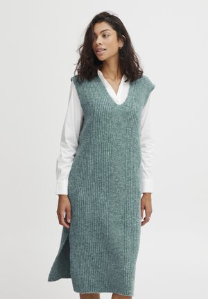 Mintgrünes gestricktes Sweatkleid mit vertikalen Rippen, V-Ausschnitt und ärmellosem Design, das seitliche Schlitze aufweist und über einem weißen Hemd getragen wird.
