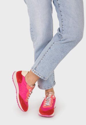 Gambe che indossano jeans azzurri chiari e sneakers rosa acceso e rosse con lacci bianchi, in posizione incrociata su sfondo bianco.