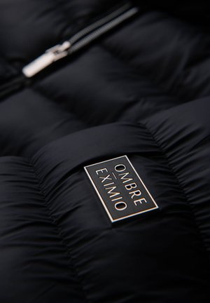 Schwarze Daunenjacke mit gestepptem Muster, ausgestattet mit einem silbernen Reißverschluss und einem rechteckigen Etikett, auf dem "OMBRA EXIMIO" in Weiß und goldenen Akzenten steht.