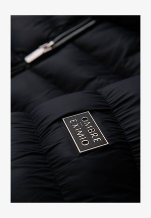 Schwarze Daunenjacke mit gestepptem Muster, ausgestattet mit einem silbernen Reißverschluss und einem rechteckigen Etikett, auf dem "OMBRA EXIMIO" in Weiß und goldenen Akzenten steht.
