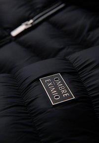 Veste matelassée noire avec une texture en quilting, dotée d'une fermeture éclair argentée et d'une étiquette rectangulaire portant "OMBRA EXIMIO" avec des accents en blanc et en or.