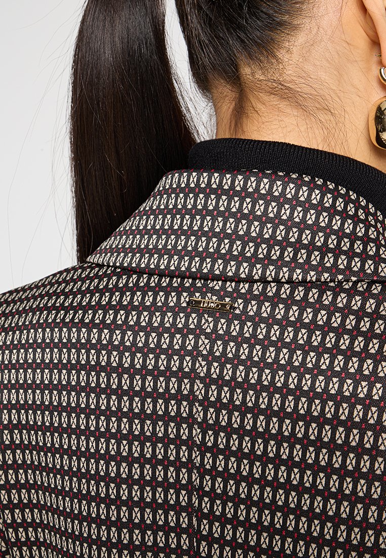 Schwarze Blazer mit geometrischem Muster und weißen sowie roten Akzenten, Kragen detail und einem goldenen Logo-Tag auf der Rückseite.