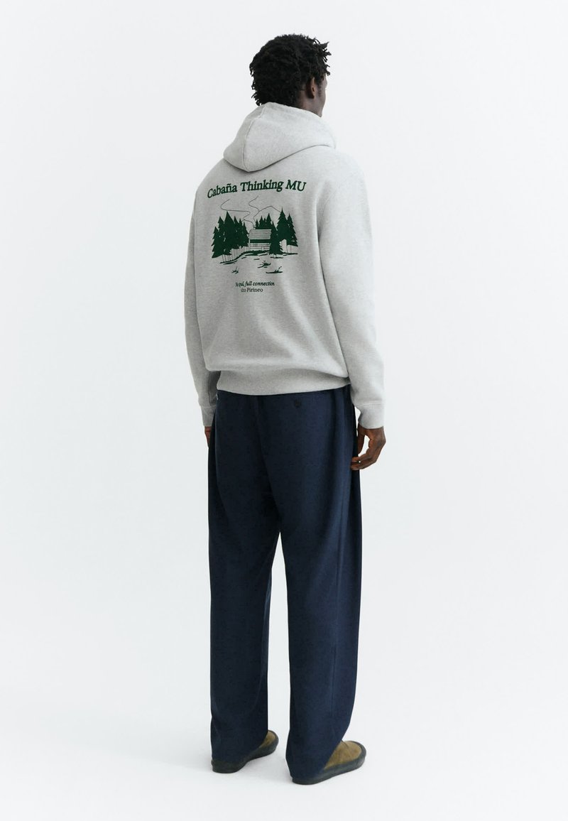 Homme de dos portant un sweat à capuche gris clair avec un motif de cabane verte et d’arbres et un pantalon ample foncé sur fond blanc.