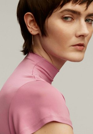 Top ajustado de color rosa con cuello alto hecho de material suave. Manga corta, diseño minimalista. La modelo tiene el pelo corto y una arete delicado.