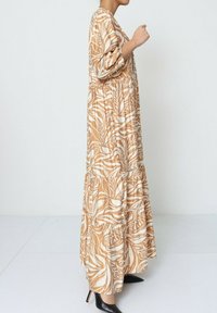 Robe longue à motifs en beige et blanc, avec des motifs floraux, taille élastique et jupe à volants. Encolure ronde et manches bouffantes.