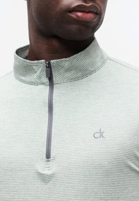 Κοντινό πλάνο ενός ατόμου που φορά ένα ανοιχτοπράσινο ριγέ πουκάμισο με φερμουάρ και ψεύτικο λαιμόκοψη, με το λογότυπο "ck" στο στήθος, εμφανίζονται τα χείλη και το κάτω μέρος του προσώπου.