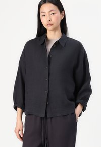Black linen button-up shirt with a collar, dropped shoulders, and rolled sleeves. Features brown buttons and a relaxed fit.