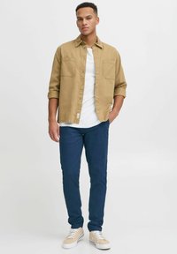 Camisa de botones marrón claro sobre una camiseta blanca, combinada con pantalones azul marino y zapatillas beige. La camisa tiene dos bolsillos en el pecho.